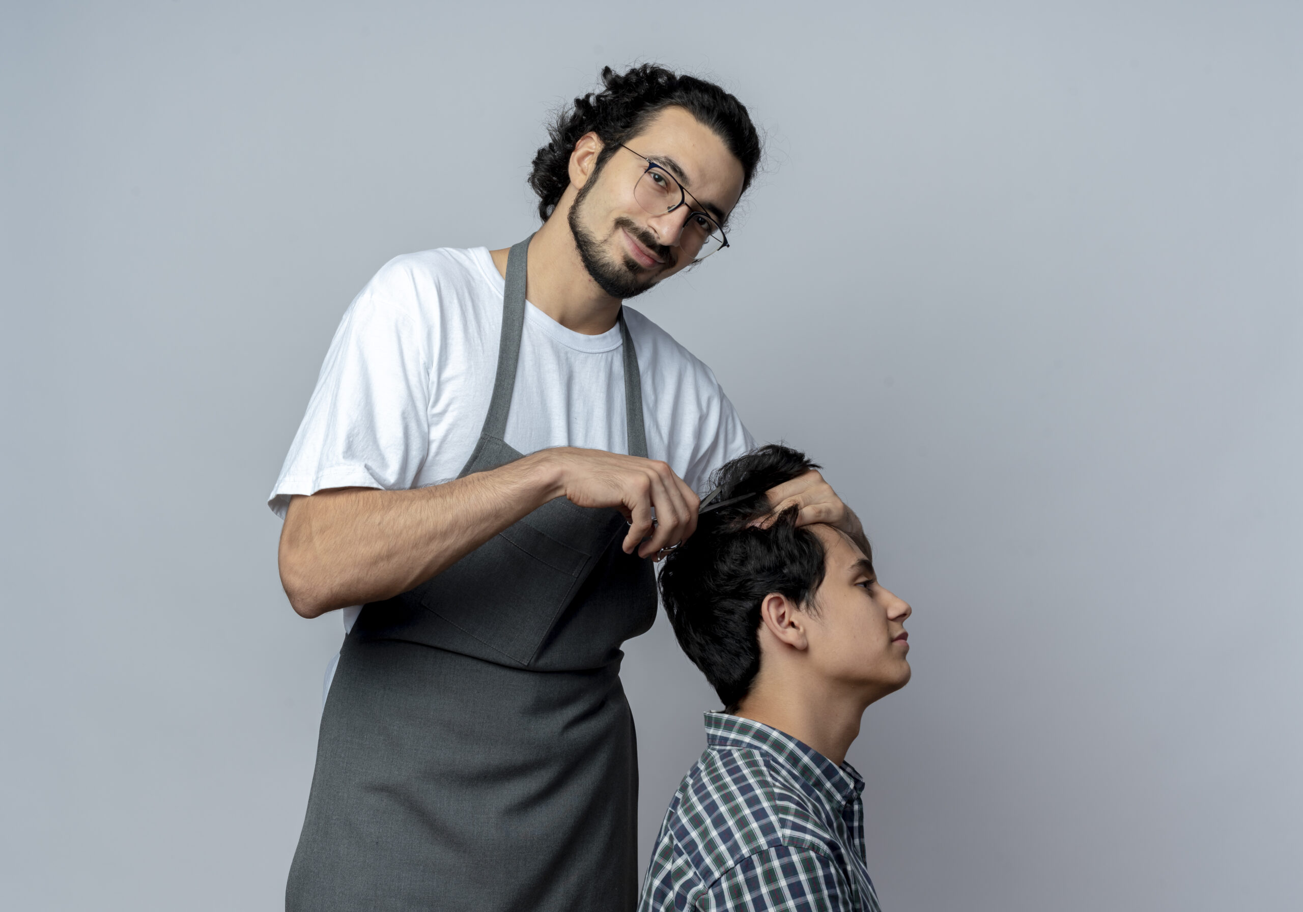 pleased young caucasian male barber wearing glasses wavy hair band uniform doing haircut his young client scaled